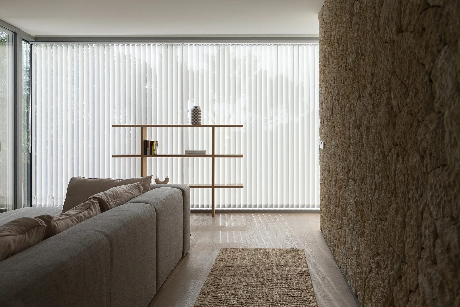 Lamas verticales blancas en salón con pared de piedra natural y estantería de madera