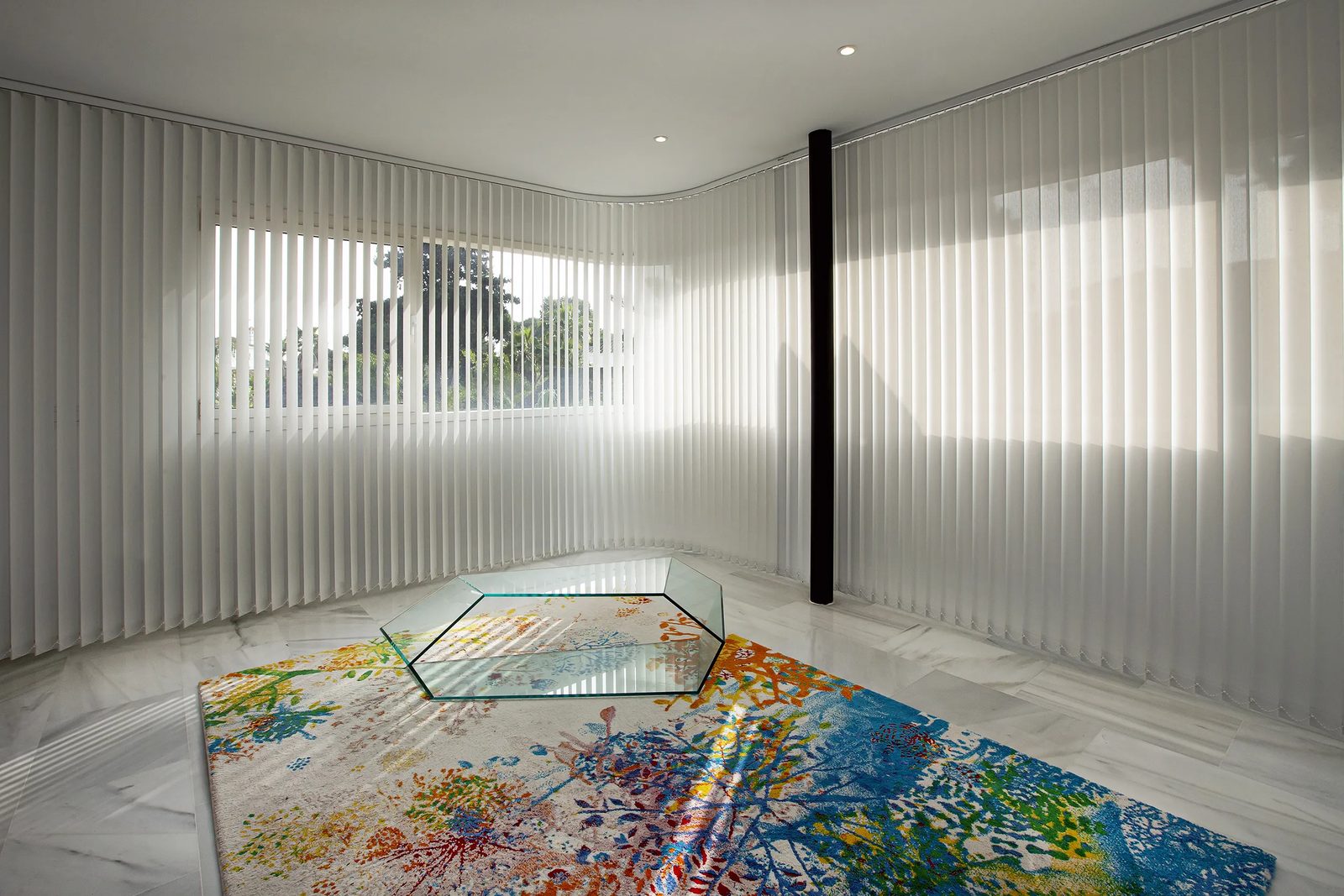Verticales blancas en sala circular con alfombra artística y mesa de cristal